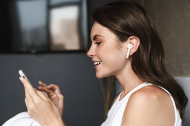 Evdeki kanepede dinlenirken kablosuz kulaklık ve akıllı telefon kullanan esmer kadın görüntüsü