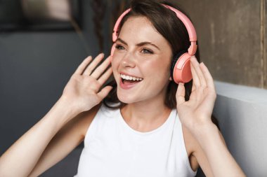 Evdeki kanepede otururken gülümseyen ve kablosuz kulaklık kullanan güzel bir kadın görüntüsü.
