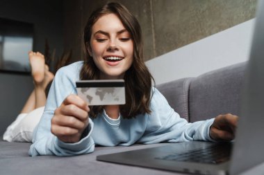 Kredi kartı tutan ve evinde kanepede yatarken dizüstü bilgisayarla çalışan neşeli bir kadın resmi.
