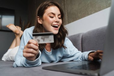 Kredi kartı tutan ve evinde kanepede yatarken dizüstü bilgisayarla çalışan neşeli bir kadın resmi.