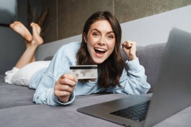 Kanepede dizüstü bilgisayarla çalışırken elinde kredi kartıyla kazanan kadının görüntüsü