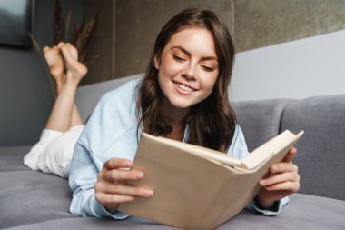 Evdeki kanepede yatarken gülümseyen ve kitap okuyan neşeli bir kadın resmi.