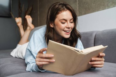 Evdeki kanepede yatarken gülümseyen ve kitap okuyan neşeli bir kadın resmi.