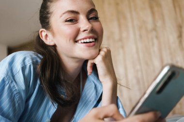 Oturma odasında yatarken gülümseyen ve cep telefonu kullanan mutlu bir kızın fotoğrafı.