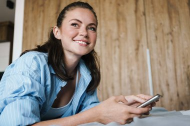 Oturma odasında yatarken gülümseyen ve cep telefonu kullanan mutlu bir kızın fotoğrafı.