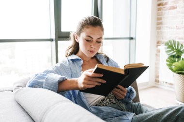 Evde kitap okurken kanepede oturan konsantre genç bir kızın görüntüsü.