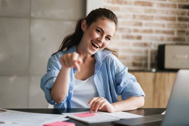 Evde dizüstü bilgisayarla notlar yazıp seni işaret eden neşeli genç bir kızın resmi.