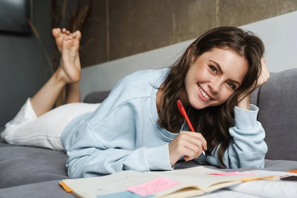 自宅でソファに横になっている間 練習帳で宿題をしている陽気な女性のイメージ — ストック写真