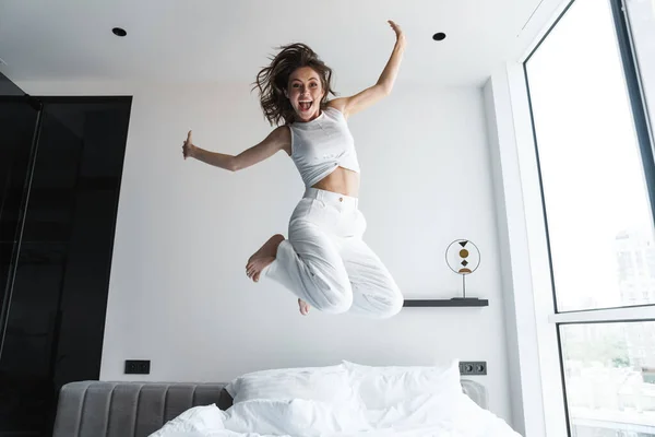 Image of young excited woman making fun and jumping on bed at home