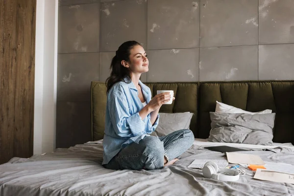 图片中 一个年轻快乐的女孩坐在室内喝咖啡 却坐在纸质文件旁边 — 图库照片