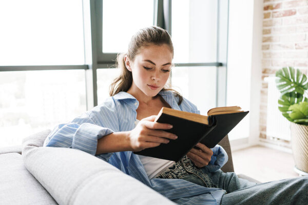 Image of a young concentrated girl indoors at home sitting on sofa while reading book