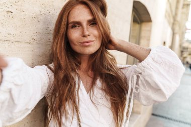 Şehirde yürürken gülümseyen ve selfie çeken neşeli kızıl kadın görüntüsü