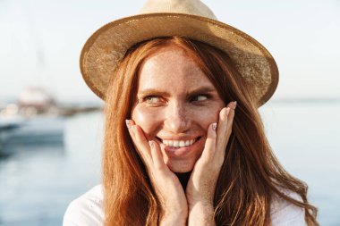 Gezintide yürürken şapkalı, gülümseyen ve yan bakan neşeli kızıl kadın görüntüsü.