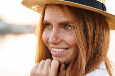 Gezinti güvertesinde yürürken gülümseyen ve yan bakan şapkalı neşeli kızıl saçlı kadın.