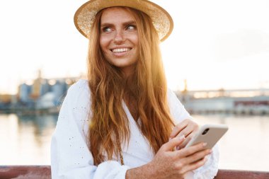 Gezintide yürürken gülümseyen ve cep telefonu kullanan neşeli kızıl kadın görüntüsü