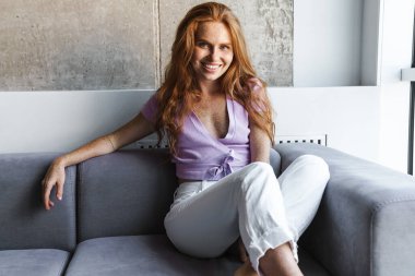 Evdeki kanepede otururken gülümseyen ve kameraya bakan kızıl saçlı kadın.