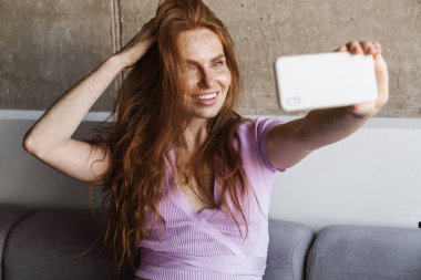Evdeki kanepede otururken cep telefonuyla selfie çeken kızıl saçlı bir kadın.