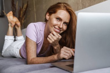 Evde kanepede yatarken gülümseyen ve laptop kullanan kızıl saçlı bir kadın resmi.