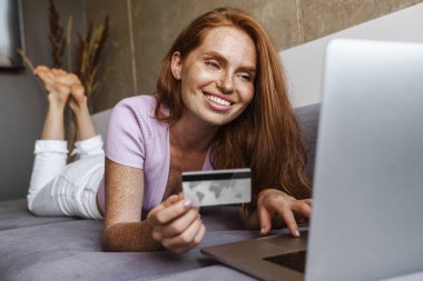 Evde kanepede yatarken elinde kredi kartı olan ve dizüstü bilgisayar kullanan kızıl saçlı bir kadın.