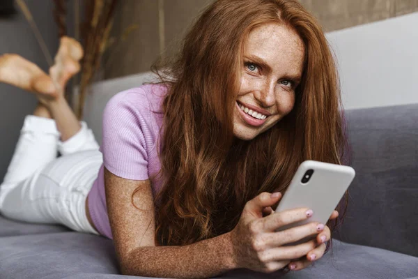 自宅でソファに横になりながら笑顔と携帯電話を使用して赤毛の幸せな女性のイメージ — ストック写真