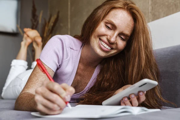 Obraz Rudej Szczęśliwej Kobiety Korzystającej Telefonu Komórkowego Zapisującej Notatki Leżąc — Zdjęcie stockowe