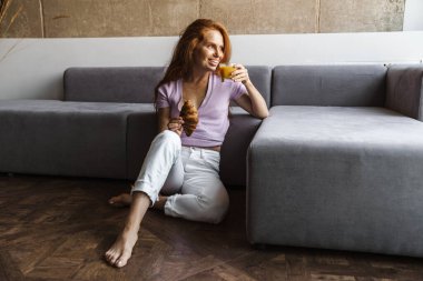 Kızıl saçlı, neşeli bir kadının evde otururken gülümseyip kahvaltı yaptığı görüntüsü.