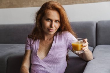Kızıl saçlı, memnun bir kadının evde oturmuş meyve suyu içişi.