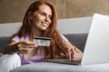 Elinde kredi kartı olan ve dizüstü bilgisayar kullanan kızıl saçlı bir kadın resmi.