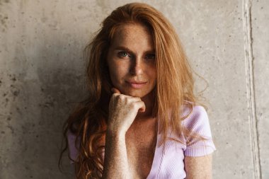 Kızıl saçlı kadının evde beton duvarın yanında poz verirken kameraya baktığı görüntü