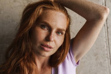 Kızıl saçlı kadının evde beton duvarın yanında poz verirken kameraya baktığı görüntü