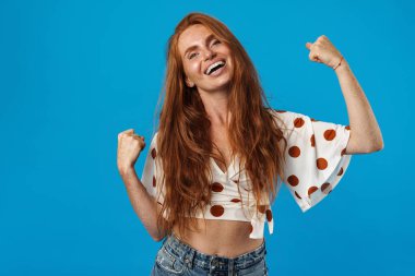 Mavi arka planda izole edilmiş başarıyı kutlayan yaz kıyafetleri giymiş mutlu genç bir kızın portresi.