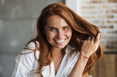 Mutlu kızıl saçlı, çilli, gülümseyen ve evde kameraya bakan bir kadın resmi.