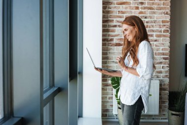 Kızıl saçlı, gülümseyen bir iş kadınının, evdeki pencerenin yanında dikilirken dizüstü bilgisayarından video konuşması.