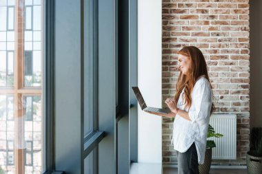 Kızıl saçlı, gülümseyen bir iş kadınının, evdeki pencerenin yanında dikilirken dizüstü bilgisayarından video konuşması.