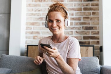 Evde kanepede otururken kulaklık ve cep telefonu kullanan gülümseyen genç bir kadın görüntüsü