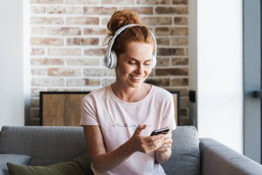 Evde kanepede otururken kulaklık ve cep telefonu kullanan gülümseyen genç bir kadın görüntüsü