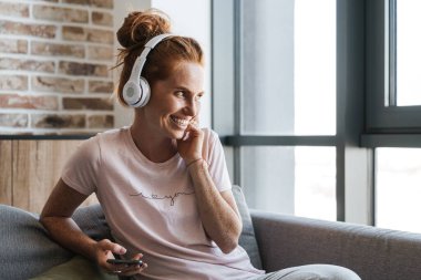 Evde kanepede otururken kulaklık ve cep telefonu kullanan gülen genç bir kadın görüntüsü