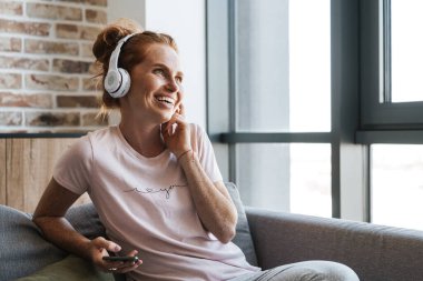 Evde kanepede otururken kulaklık ve cep telefonu kullanan gülen genç bir kadın görüntüsü