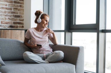 Evde kanepede otururken kulaklık ve cep telefonu kullanan gülümseyen genç bir kadın görüntüsü
