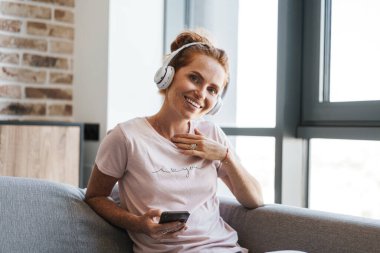 Evde kanepede otururken kulaklık ve cep telefonu kullanan gülümseyen genç bir kadın görüntüsü