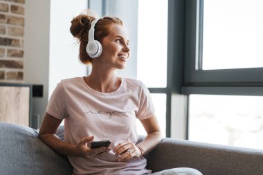 Evde kanepede otururken kulaklık ve cep telefonu kullanan gülümseyen genç bir kadın görüntüsü