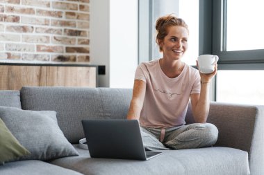 Neşeli kızıl saçlı bir kadının kahve içip dizüstü bilgisayar kullanması.