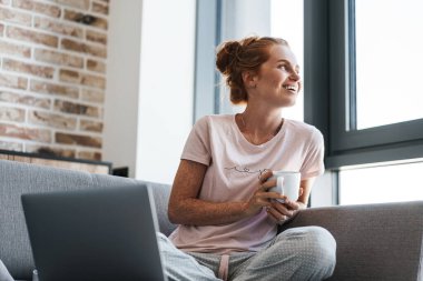 Neşeli kızıl saçlı bir kadının kahve içip dizüstü bilgisayar kullanması.