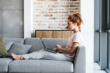 Evde kanepede otururken dizüstü bilgisayarla çalışan odaklanmış kızıl saçlı bir kadın.