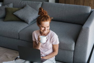 Kahvesini yudumlayan ve dizüstü bilgisayar kullanan neşeli bir kızıl kadın resmi.