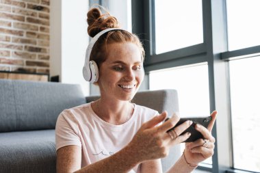 Kulaklıkla gülümseyen ve evde otururken cep telefonuyla oyun oynayan bir kadın görüntüsü