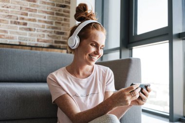 Kulaklıkla gülümseyen ve evde otururken cep telefonuyla oyun oynayan bir kadın görüntüsü