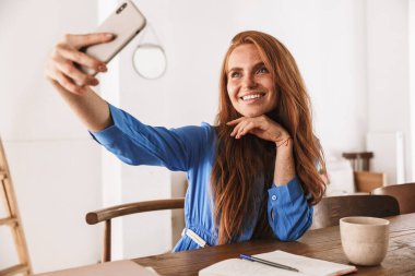 Kafede dinlenirken ve bir fincan kahve içerken akıllı telefonuyla selfie çeken güzel, genç bir kadın.