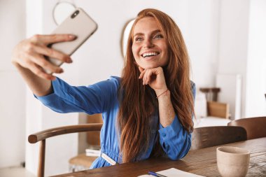 Kafede dinlenirken ve bir fincan kahve içerken akıllı telefonuyla selfie çeken güzel, genç bir kadın.
