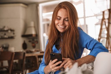 Mutfakta rahat bir şekilde otururken gülümseyen ve cep telefonu kullanan mutlu kızıl kadın görüntüsü